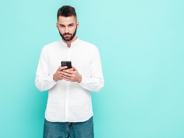 Hübsches, selbstbewusstes Modell, sexy, stilvoller Mann in Hemd und Jeans, modischer Hipster-Mann, der in der Nähe der blauen Wand im Studio posiert und Smartphone hält, Handy-Bildschirm mit Apps betrachtet