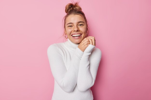 Hüftaufnahme einer glücklichen, begeisterten europäischen Frau, die breit lächelt, hält die Hände zusammen und fühlt sich froh, Freizeit in Gesellschaft von Freunden zu verbringen, trägt weiße Rollkragen-Posen vor rosa Hintergrund.