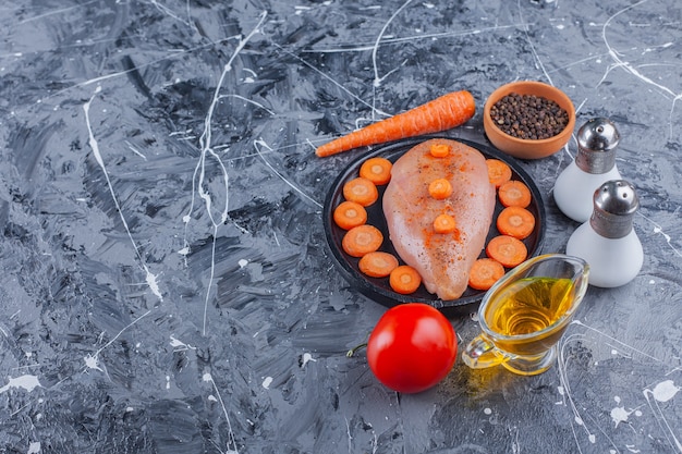 Hühnerbrust und geschnittene Karotten auf einem Teller neben Salz, Öl, Gewürzen, Karotten und Tomaten auf der blauen Oberfläche