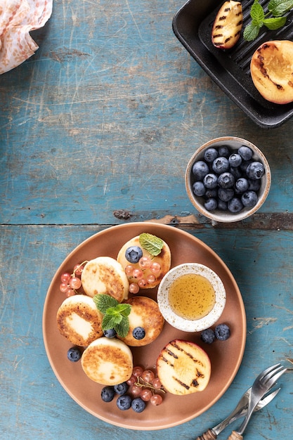 Kostenloses Foto hüttenkäse-pfannkuchen käsekuchen ricotta krapfen mit frischen heidelbeeren, johannisbeeren und pfirsiche auf einem teller gesundes und leckeres frühstück für den urlaub blauer hintergrund aus holz