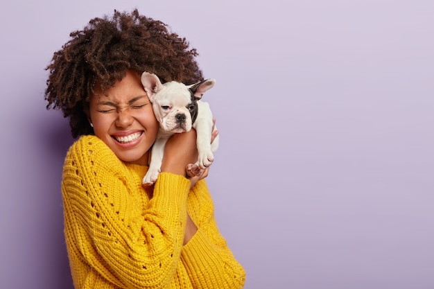 Hundebesitzerin und ihr Haustier. Glückliches ethnisches lockiges Mädchen hält niedlichen kleinen Welpen nahe Gesicht, drückt Liebe und Fürsorge zum Haustier aus, kauft Hund der Lieblingsrasse, lacht, hat Augen vor Vergnügen geschlossen