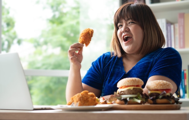 Hungrige übergewichtige frau mit fried chicken hamburger auf einem holzteller und pizza auf dem tisch