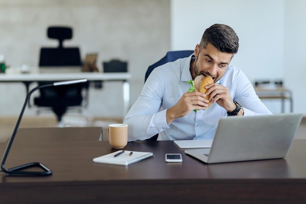 Hungriger Geschäftsmann, der Sandwich isst, während er Laptop im Büro benutzt