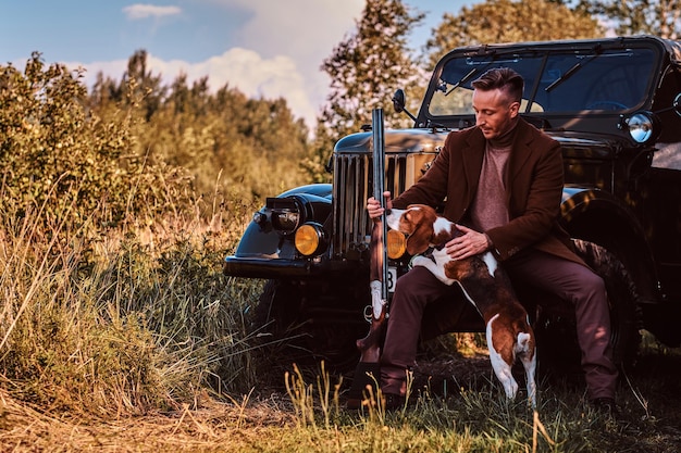 Hunter in eleganter Kleidung hält eine Schrotflinte und sitzt zusammen mit seinem Beagle-Hund, während er sich in einem Wald an ein Retro-Militärauto lehnt.