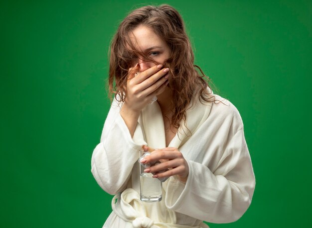Husten junges krankes Mädchen, das weißes Gewand hält Glas des Wassers mit Pillen und bedecktem Mund mit Hand lokalisiert auf Grün