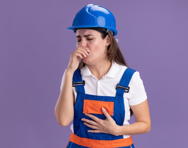 Hustende junge Baumeisterfrauen in Uniform, die Hand auf den Mund legen, isoliert auf lila Wand