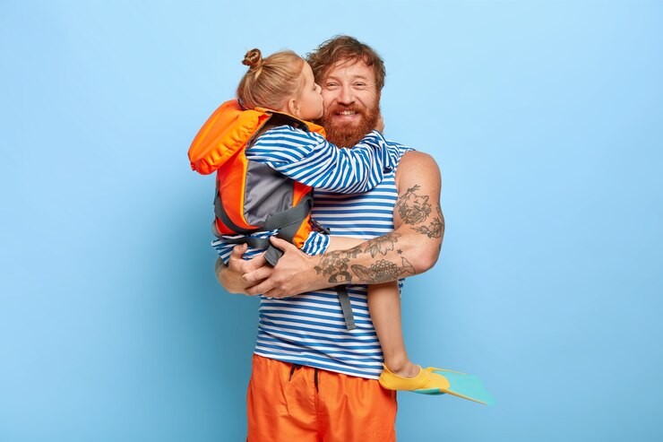 Kostenlose Foto ich bin froh, dass mein vater glücklich ist, einen liebevollen kuss und eine umarmung von der tochter zu bekommen, hält sie an den händen, trägt matrosenpullover, trägt eine orangefarbene schwimmweste und flossen, verbringt die sommerferien zusammen und hat spaß