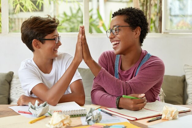 Ich bin froh, dass multiethnische dunkelhäutige Frauen und Jugendliche sich gegenseitig High Five geben, am Arbeitsplatz sitzen, gute Ergebnisse erzielen, während sie zusammen studieren, Aufzeichnungen in den Notizblock schreiben und ihre Zustimmung demonstrieren