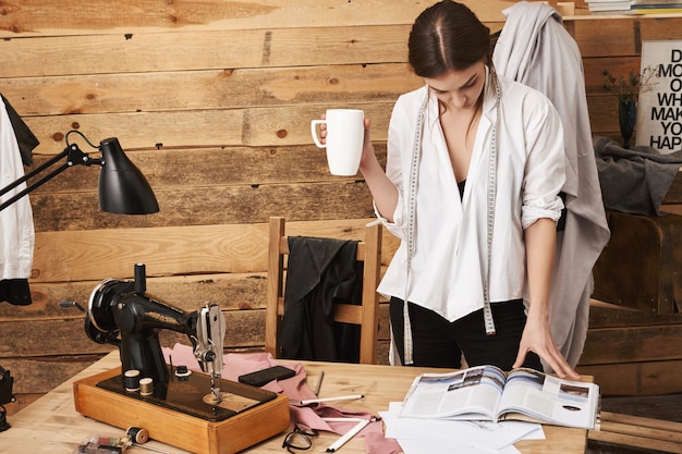 Kostenloses Foto ich brauche inspiration. porträt des niedlichen weiblichen schneiderlesemagazins, während tee trinkend und pause in der werkstatt, mit nähmaschine unter neuem kleidungsstück arbeitend