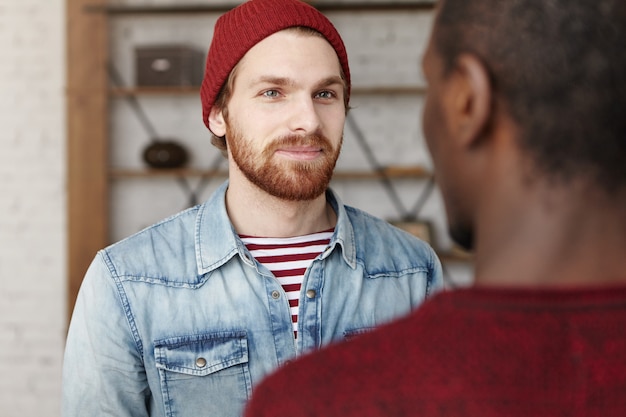 Kostenloses Foto ich freue mich für dich. innenaufnahme eines hübschen modischen jungen kaukasischen mannes, der mit seinem afroamerikanischen schulkameraden spricht, den er seit ewigkeiten nicht mehr gesehen hat. zwei freunde, die sich nett unterhalten und über neuigkeiten diskutieren