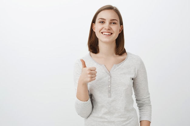 Ich gebe Idee Daumen hoch. Erfreut glückliche charmante junge europäische Frau mit kurzen braunen Haaren, die mit fröhlichem Ausdruck lächeln und guten Blick des Freundes mögen, während sie neuen Outfir anprobieren, der über graue Wand genehmigt