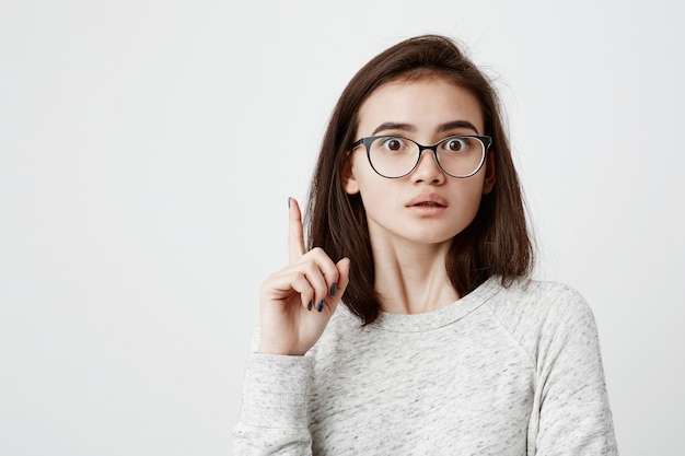 Ich habe eine großartige Idee. Kluge, schlagfertige, hübsche Frau mit dunklem Haar und Brille, die den Finger nach oben zeigt. Netter weiblicher Teenager mit geknallten Augen, Geste mit Zeigefinger machend