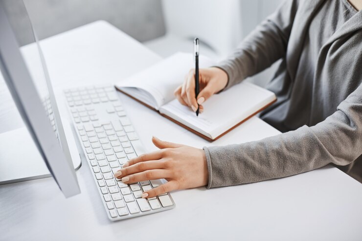 Kostenlose Foto ich kann mehrere aufgaben erledigen. beschnittenes schuss erfolgreiches mädchen, das auf tastatur tippt und notizen macht, während auf computerbildschirm betrachtet und neue geschäftsgrafik studiert. es ist keine zeit für ruhe