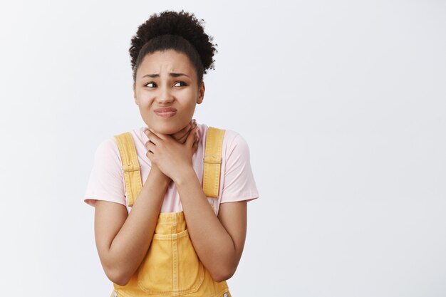 Ich kann nicht atmen, hilf mir. Porträt eines unzufriedenen unglücklichen afroamerikanischen Mädchens in gelben Overalls, Händchen haltend am Hals, Würgen oder Halsgefühl, Unbehagen und richtiges Starren