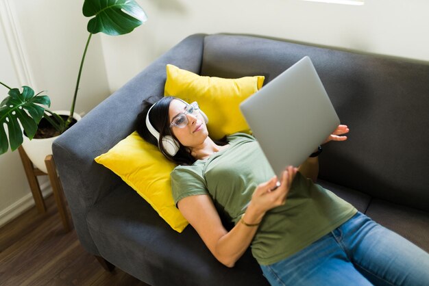 Ich liebe es, zu Hause zu lernen. Entspannte junge Frau, die sich auf die Couch legt, während sie einer virtuellen Universitätsklasse zuhört