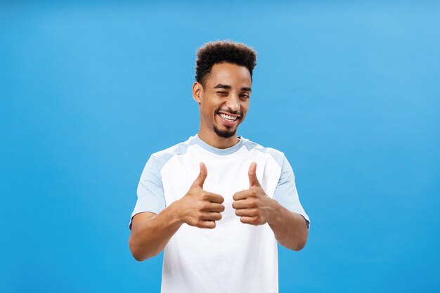 Ihr macht das super, unterstützt euch. Zufriedener kreativer und stilvoller junger afroamerikanischer Mann mit Bart, der freudig lächelt und breit Daumen hoch zeigt oder wie Geste über blauer Wand.
