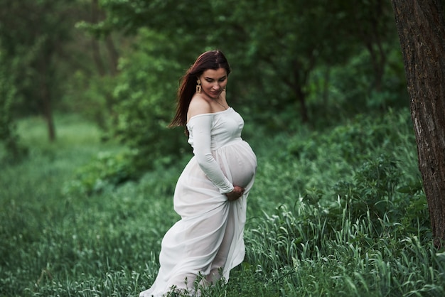 Im Gras stehen. Schöne schwangere Frau im Kleid haben einen Spaziergang im Freien. Positive Brünette