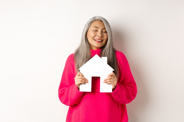 Immobilie. Schöne verträumte asiatische Dame, die das Papierhausmodell mit geschlossenen Augen umarmt, lächelt, als träumt sie davon, eine Wohnung zu kaufen, steht auf weißem Hintergrund