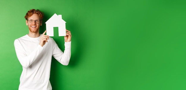 Immobilien hübscher junger Mann mit roten Haaren, der eine Brille trägt, die einen Papierhausausschnitt zeigt und lächelt