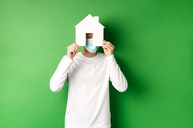 Immobilien- und Covid-19-Pandemiekonzept. Lustiger junger Mann in Gesichtsmaske und weißem Langarm-Versteckgesicht hinter Papierhausausschnitt, Blick in die Kamera, grüner Hintergrund