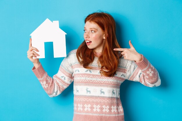 Immobilienkonzept. Aufgeregte rothaarige Frau mit roten Haaren, die auf das Papierhausmodell zeigt und es betrachtet, die Wohnungswerbung zeigt und auf blauem Hintergrund steht.