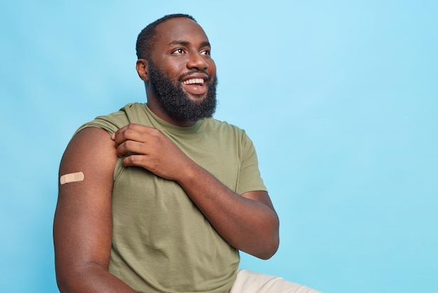Impfkampagne gegen Covid 19. Fröhlicher dunkelhäutiger Mann zeigt nach der Impfung einen Arm mit Klebepflaster, erhält Impfstoff-Posen gegen den blauen Kopierbereich für Ihren Text
