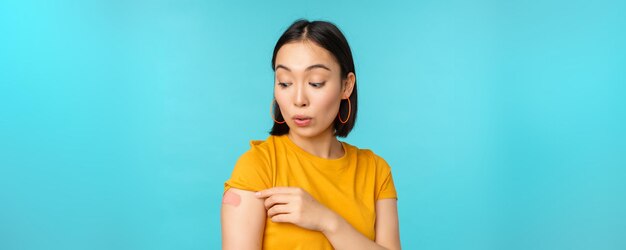 Impfkampagne von covid19 Junge, schöne, gesunde asiatin, die Schulter mit Bandaid-Impfkonzept zeigt, das über blauem Hintergrund steht