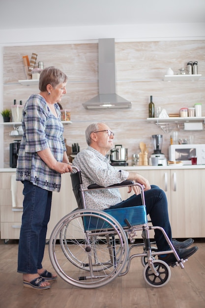 Kostenloses Foto in der küche schaut eine alte frau auf einen behinderten ehemann, der im rollstuhl sitzt. behinderter älterer mann, der im rollstuhl in der küche sitzt und durch das fenster schaut. leben mit behinderten. ehefrau hilft