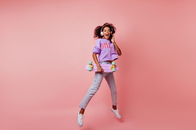 In voller Länge Aufnahme einer atemberaubenden schwarzen Frau in stilvollen Jeans, die auf Rosa springen. lockiges afrikanisches Mädchen mit Skateboard, das positive Emotionen ausdrückt.