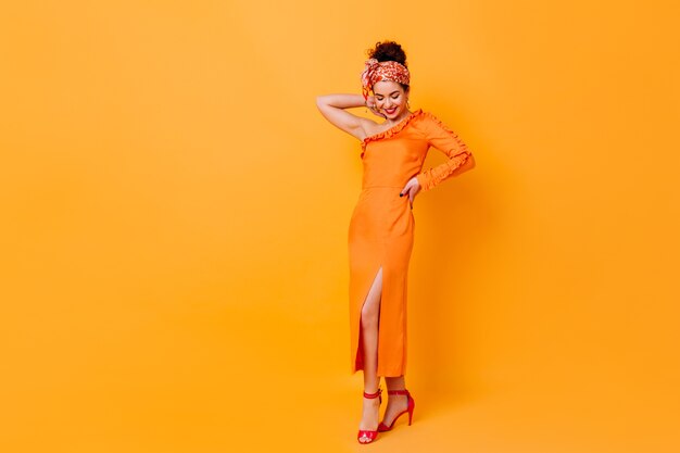 In voller Länge Aufnahme einer modischen Dame in einem langen Seidenkleid mit Schlitz und Schal über dem Kopf, die auf orangefarbenem Raum posiert.