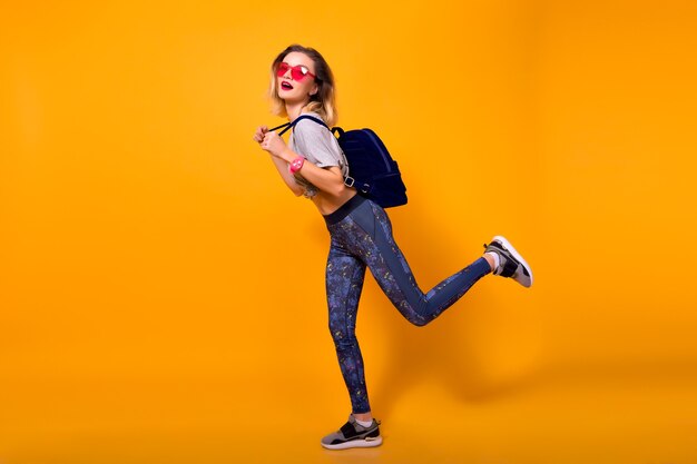 In voller Länge Innenporträt des Mädchens, das Leggings trägt, läuft auf gelbem Hintergrund. Ziemlich schlankes weibliches Model in Turnschuhen mit Rucksack, der im Studio herumalbert.