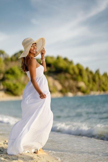 In voller Länge von einer glücklichen Frau im weißen Kleid, die während des Sommertages am Sandstrand steht