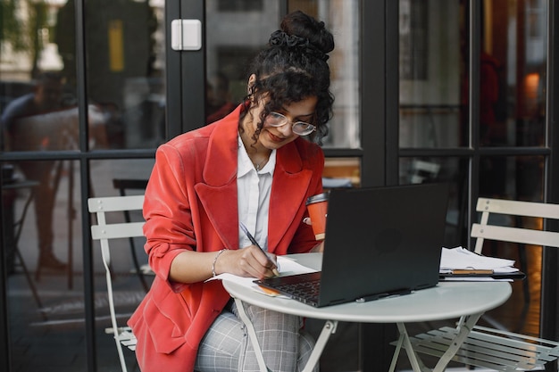 Indische Frau arbeitet an einem Laptop in einem Straßencafé. Stylische schicke Kleidung tragen - Jacke, Brille