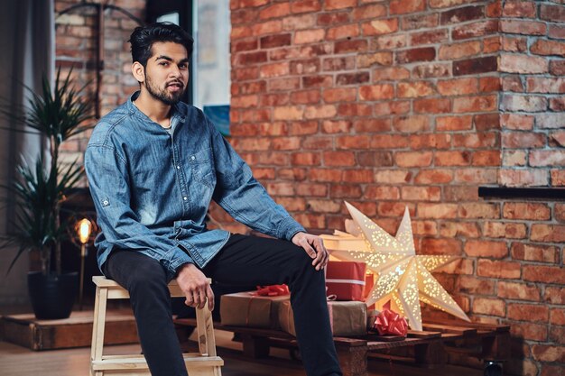 Indischer bärtiger Hipster-Mann mit Tätowierungen auf dem Arm in einem Loft-Zimmer mit Weihnachtsdekoration.