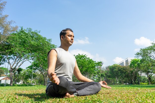 Indischer Mann, der draußen in der Lotoshaltung auf Sommerrasen mit Bäumen im Hintergrund meditiert.