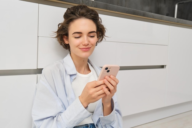 Indoor-Aufnahme eines glücklichen Mädchens mit Smartphone sitzt zu Hause auf dem Boden und nutzt das Handy, um Video zu beobachten