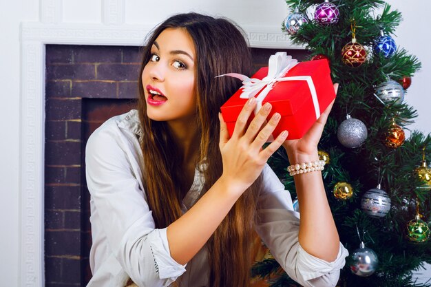 Indoor-Lifestyle-Porträt eines hübschen lächelnden Mädchens mit hellem Make-up und erstaunlichen brünetten langen Haaren