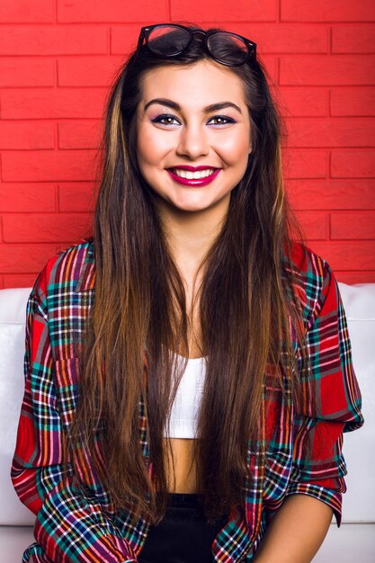 Indoor-Mode-Lifestyle-Porträt einer jungen hübschen Hipster-Frau mit hellem Make-up und erstaunlichem Lächeln, haben lange brünette Haare und tragen kariertes Hemd. Rote Stadtmauer.