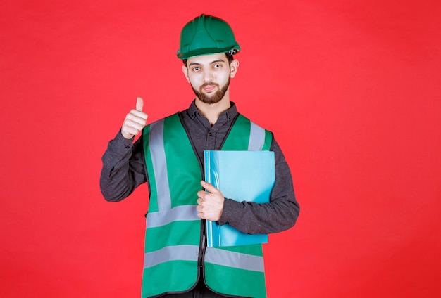 Ingenieur in grüner Uniform und Helm, der einen blauen Ordner hält und ein positives Handzeichen zeigt.