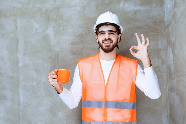 Ingenieur Mann im weißen Helm hält einen orangefarbenen Becher und genießt den Geschmack des Getränks.