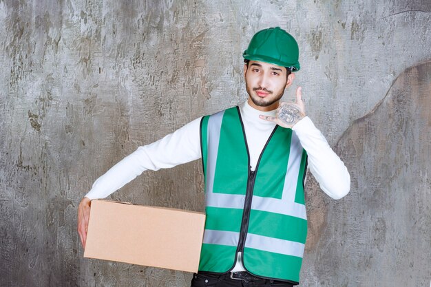 Ingenieur Mann in gelber Uniform und Helm hält ein Papppaket und bittet um einen Anruf.