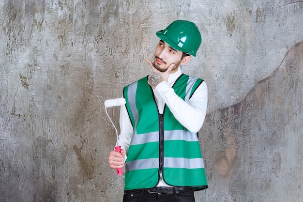 Ingenieur Mann in gelber Uniform und Helm hält eine Trimmrolle für Wandmalerei und sieht nachdenklich aus.