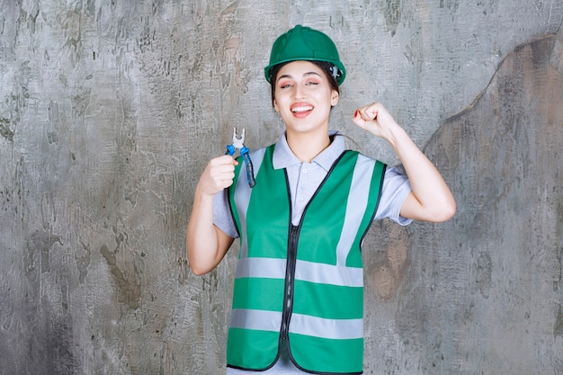 Ingenieurin im grünen Helm, der eine Zange für Reparaturarbeiten hält und ihre Faust zeigt.