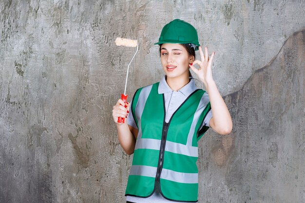 Ingenieurin im grünen Helm, die eine Trimmrolle für die Wandmalerei hält und ein positives Handzeichen zeigt.