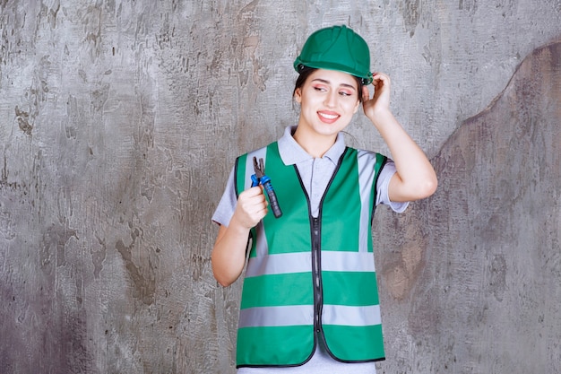 Ingenieurin im grünen Helm hält Zange für Reparaturarbeiten und sieht verwirrt und nachdenklich aus.