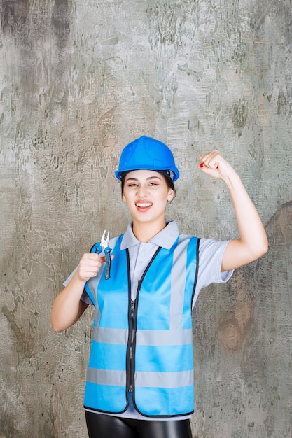 Ingenieurin in blauer ausrüstung und helm, die eine zange für reparaturarbeiten hält und ihre faust zeigt. Premium Fotos