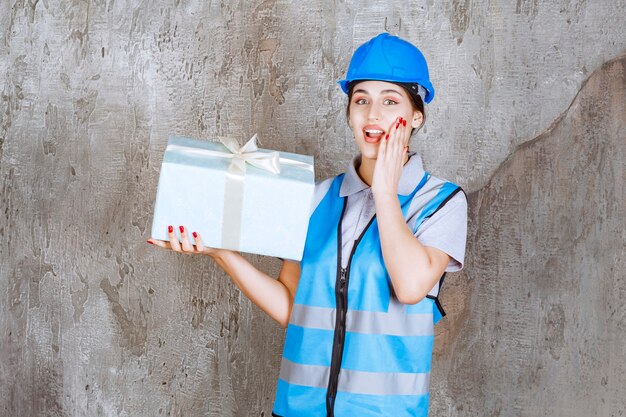 Ingenieurin in blauer Uniform und Helm, die eine blaue Geschenkbox hält und überrascht und aufgeregt aussieht