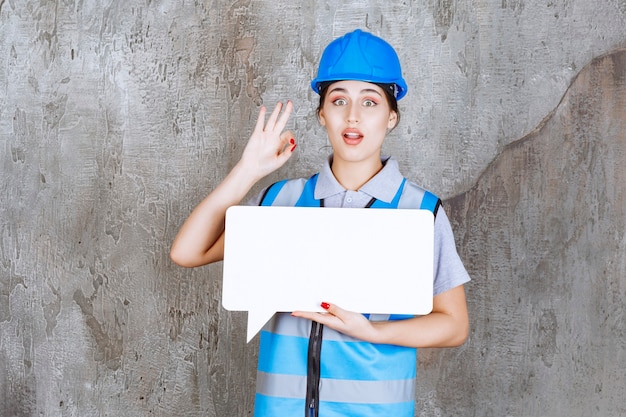 Kostenloses Foto ingenieurin in blauer uniform und helm, die eine leere rechteck-infotafel hält und ein genusszeichen zeigt.