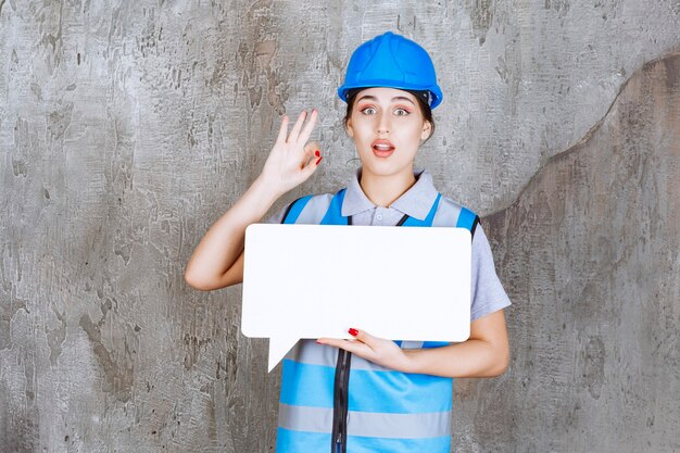 Ingenieurin in blauer Uniform und Helm, die eine leere Rechteck-Infotafel hält und ein Genusszeichen zeigt.