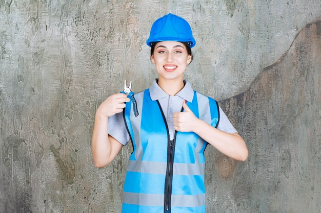 Ingenieurin in blauer Uniform und Helm, die eine Metallzange zur Reparatur hält und ein positives Handzeichen zeigt